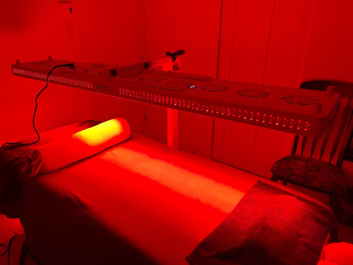 Red light therapy setup: a long LED panel above a massage table with a pillow and towels in a treatment room